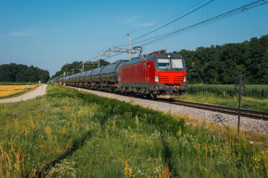 Kırsal alanda silindirik tank vagonları taşıyan kırmızı yük treni. Güneşli bir gün açık gökyüzü, yeşil tarlalar, kır çiçekleri ve rayların yanında toprak bir patika..