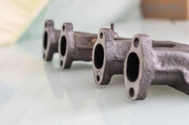 Paslı dökme demir egzoz manifoldu, yeni makinelenmiş, düzleştirilmiş iskele yüzleri, parlak beyaz bir yüzey üzerinde görüntüleme veya teftiş için yatıyor.