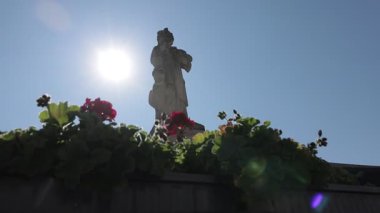 Obernberg Marktplatz 'da berrak mavi gökyüzünde parlak bir güneşe karşı silueti olan cüppeli bir heykelin heykeli dimdik ayakta duruyor. Heykel, canlı kırmızı çiçekler ve ahşap bir çıkıntının üzerindeki yemyeşil yapraklar tarafından çerçevelenmiş ve pitoresk bir manzara yaratıyor..