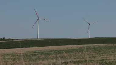Kırmızı uçlu bıçakları olan modern rüzgar türbinleri açık mavi gökyüzünün altındaki çimenli bir tepenin üzerinde dimdik dururlar. Manzara, yenilenebilir enerji ve kırsal manzaranın bir karışımını gösteren, uzak bir ağaç hattı ile geniş yeşil ve kahverengi alanlar içerir..