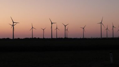 Yumuşak pembe ve mor bir akşam gökyüzüne karşı bir sıra rüzgar türbini duruyor. Aşağıdaki karanlık manzarada tarlalar ve uzak ağaçlar, günbatımının yumuşak parlaklığıyla dingin bir zıtlık yaratarak yenilenebilir enerjiyi vurguluyor.