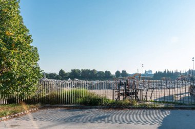 Tadilat sırasında Ljubljana 'daki bir spor merkezinde yıkım alanı. Beton tribünler kısmen yıkıldı, kazıcılar ve çöp kamyonları açık mavi gökyüzünün altında dağınık bulutlarla kırmızı ve sarı enkazı temizliyor. Ağaçları çevreleyen