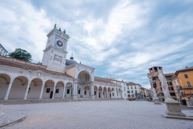 Udine, İtalya 'daki Iconic Piazza della Libert' te Loggia del Lionello ve Clock Tower sergilenmektedir. Venedik Gotik mimarisi, kemerler, merdivenler ve bulutlu gökyüzünün altında bisikletçilerle dolu canlı bir meydan..