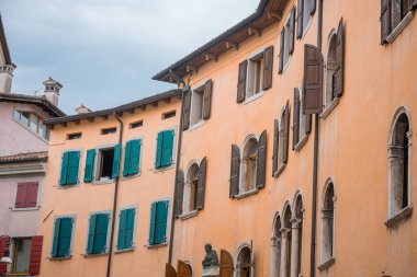 İtalya 'nın Udine şehrinde şeftali rengi duvarları, kemerli pencereleri, deniz mavisi panjurları ve bulutlu gökyüzüne karşı çerçevelenmiş bir heykeli olan tarihi bir binanın büyüleyici manzarası..