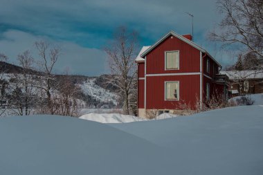İsveç 'in karlı bölgelerinde beyaz tepeler ve çıplak kış ağaçlarıyla çevrili büyüleyici kırmızı ahşap bir ev duruyor. Çam ağaçlarıyla kaplı dağlar ve mavi bir gökyüzü barışçıl bir İskandinav kış manzarası yaratır..