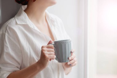 Closeup of female hands with a mug of beverage. Beautiful girl in grey sweater holding cup of tea or coffee in the morning sunlight.