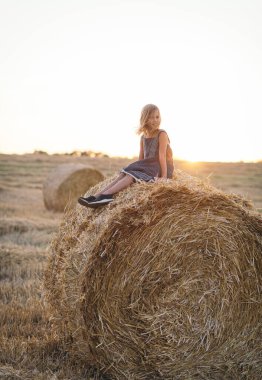 Sevimli beyaz kız çiftliğin yakınındaki buğday tarlasında altın saman balyasının üstünde oturarak eğleniyor. Mutlu çocukluk ve özgürlük kavramı. Kırsal alan manzarası