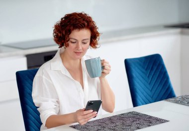 Genç bayan çay içiyor ve işe gitmek için hazırlanırken mutfağında akıllı telefon kullanıyor. Sabah evde eğlenirken arkadaşlarınla mesajlaşıyorsun.