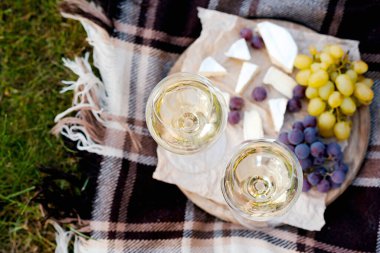 İki kadeh beyaz şarap ve dışarıda servis edilen peynirli ve üzümlü ahşap bir tabak.