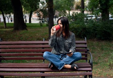 Parkta cep telefonu kullanan ve kahve içen bir kız. Sonbahar günü