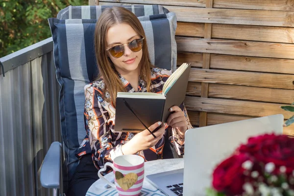 Balkonda oturup kitap ya da defter okuyan genç bir kadın. Ön planda çiçekler var. Açık hava sahnesi.