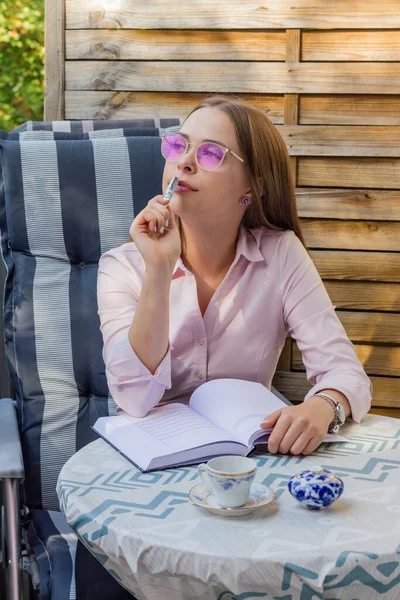Not defterine not alırken düşünen pembe gözlüklü hoş bir kızın portresi. Dünyaya gül renkli gözlüklerle bakmak. Balkonda. Öğrenme kavramı.