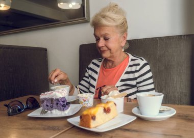 Çekici, neşeli yaşlı kadın bir kafede otururken kahve içiyor. Kafeteryadaki olgun kadın portresi. Masanın üzerinde renkli kekler ve kahve var. Rahatlatıcı bir atmosfer. Kahveli orta yaşlı güzel bir kadın..