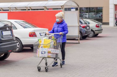 Coronavirus salgını sırasında otoparkta alışveriş arabasıyla koruyucu maske takan olgun bir kadının fotoğrafı. Günlük hayat, boşluğu kopyala. 