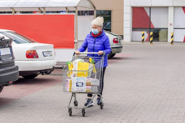 Coronavirus salgını sırasında otoparkta alışveriş arabasıyla koruyucu maske takan olgun bir kadının fotoğrafı. Günlük hayat, boşluğu kopyala. 