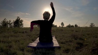 Yaşlı bir kadın batan güneşin arka planında yoga yapıyor. Huzurlu yaşlı kadın çayırda joga yapıyor. Güneş egzersiz figürüne nüfuz eder..   