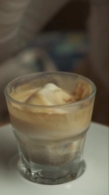 Zengin bir macchiato 'nun köpüklü tabakalarını karıştıran bir kaşığın şeffaf bir bardakta tutulduğu, dokularını gösteren ve içeceğin sıcaklığını davet eden detaylı bir video.. 