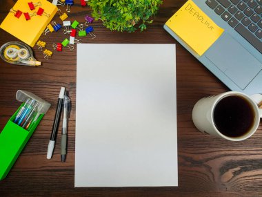 Düz uzan, sahte kağıt yap. Çalışma alanı ofis masasının arka planında, yukarıdan görülüyor. Boş beyaz kağıtlar, dizüstü bilgisayarlar, ofis malzemeleri, kalemler, yeşil yapraklar ve ahşap bir masanın arka planına karşı kahve fincanları olan bir çalışma alanı..