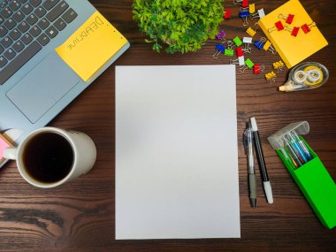 Düz uzan, sahte kağıt yap. Çalışma alanı ofis masasının arka planında, yukarıdan görülüyor. Boş beyaz kağıtlar, dizüstü bilgisayarlar, ofis malzemeleri, kalemler, yeşil yapraklar ve ahşap bir masanın arka planına karşı kahve fincanları olan bir çalışma alanı..