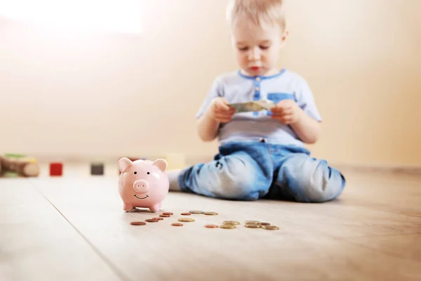 Cute boy sitting on the floor indoor and inserting euro coins in piggybank. Concept of the economy and family budget.