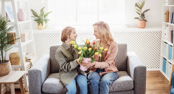 Mother and daughter sitting on the sofa with tulips in their hands indoor. Concept of the mothers day, birthday and motherhood.