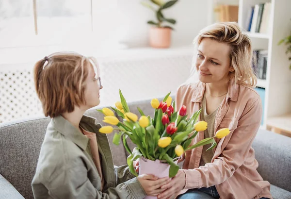Mother and daughter sitting on the sofa with tulips in their hands indoor. Concept of the mothers day, birthday and motherhood.