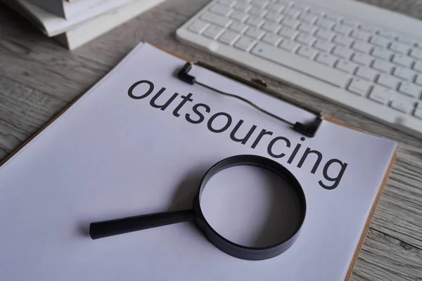 Closeup image of magnifying glass and paper clipboard with text OUTSOURCING.