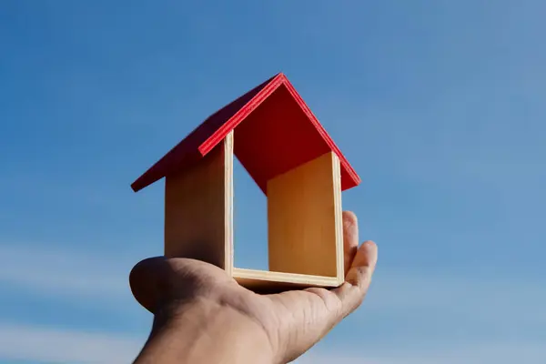 Hand holding toy wooden house with background of blue sky. Dream house, home ownership concept.