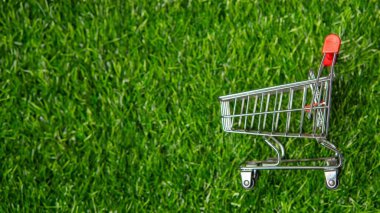 Canlı yeşil çimenler üzerinde minyatür bir alışveriş arabası kopyalama alanı sürdürülebilir alışverişi sembolize eder. Çevre dostu tüketicilik, çevre bilinci ve sorumlu satın alma kavramını vurguluyor.