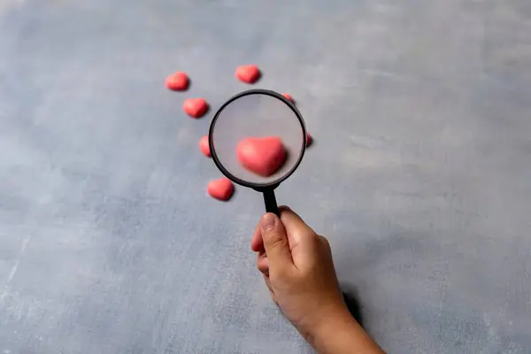 Büyüteç tutan bir el, diğerleri arasında tek bir pembe kalbe odaklanır. Aşk konsepti arayışı.