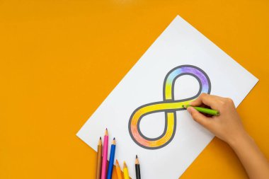 Renkli kalemlerin etrafa saçıldığı beyaz bir kağıt parçasına gökkuşağı sembolünü boyayan bir el. Otizm farkındalık kavramı.