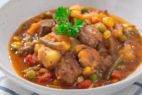 Vegetable beef stew with potato, green beans, carrot, celery, peas and corn, in gray bowl, horizontal, closeup