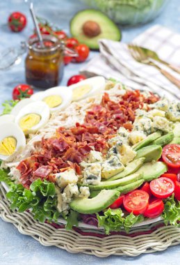 Cobb salad with greens, tomato, avocado, blue cheese, bacon, rotisserie chicken and boiled eggs, with vinaigrette sauce, on a white plate, vertical