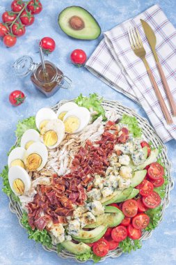 Cobb salad with greens, tomato, avocado, blue cheese, bacon, rotisserie chicken and boiled eggs, with vinaigrette sauce, on a white plate, vertical top view