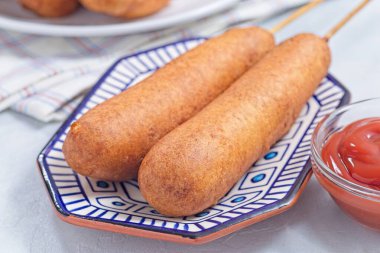 İki mısırlı sosis, mavi bir tabakta, ketçapla servis edilmiş, yatay.