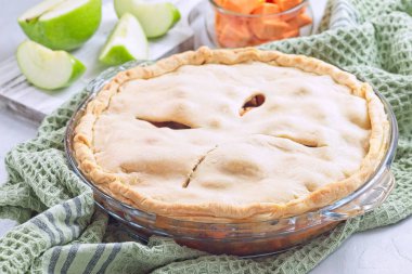 Tatlı patates ve elmalı turta cam tabakta pişiyor, taze elma dilimleri ve mutfak havlusu sıcacık sonbahar manzarası yaratıyor, yatay.