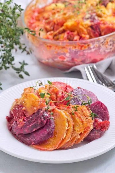 Fırında pişmiş tatlı patates, havuç ve pancar kökü graten, taze kekik ile baharatlı ve eritilmiş peynirle kaplı, beyaz bir tabakta servis ediliyor, dikey olarak.