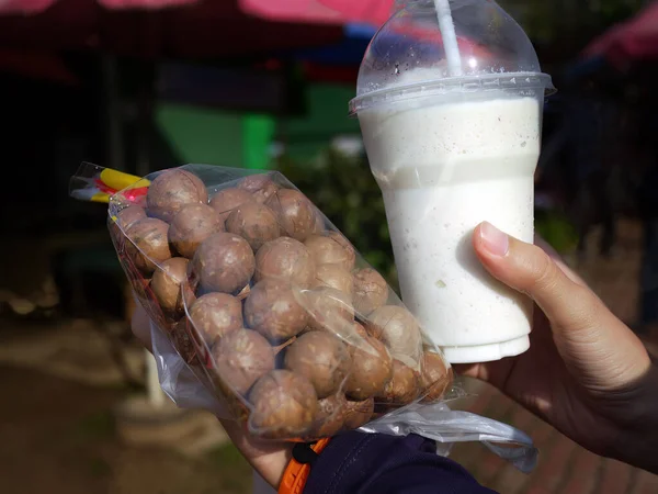 stock image Left hand holding a bag of in shell raw macadamia nuts, right hand holding a plastic cup of macadamia milk, cold drink, Dairy free milk, vegan