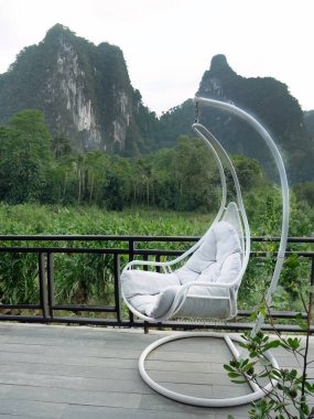 Beyaz yumurta şekilli sallanan sandalye, beyaz yastıklı, ahşap terasta salıncak dağlar ve arka planda yeşil orman manzarası