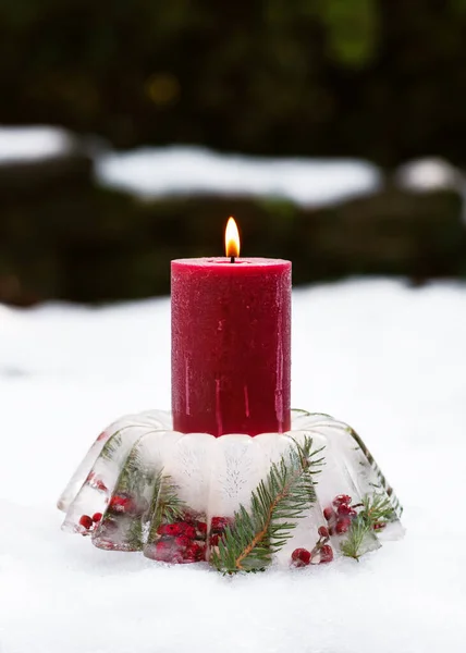 Handmade Christmas candle holder in shape of cake made of ice, red rose hips berries, holly berry branches and thuja twigs with burning candles in snow. Winter outdoor decoration concept.
