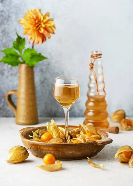 Homemade wine or liquor of golden berries in a small glass and ripe fruits on a plate. Homemade strong alcoholic beverage. Rustic style. (Physalis pubescens)