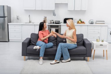 Smiling interracial friends holding glasses of champagne in kitchen  clipart