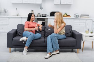 Asian woman holding champagne while talking to friend on couch at home  clipart