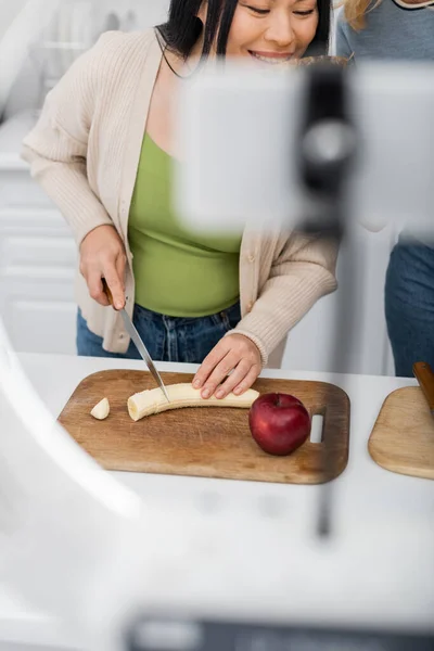 stock image Smiling asian woman cutting banana near friend and blurred smartphone 