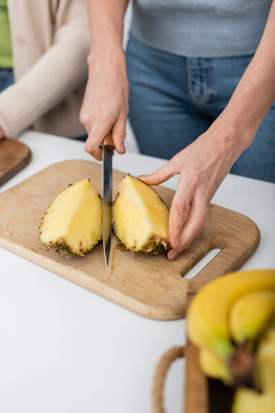 Cropped view of woman cutting fresh pineapple near blurred friend at home 