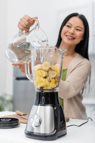 stock image Blurred asian woman pouring water in blender with fruits in kitchen 