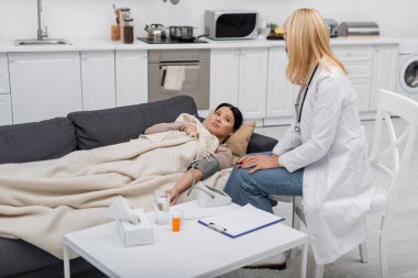 Hasta Asyalı hasta, evde sağlık maskesi ve tonometre takmış bir doktorun yanında yatıyor. 