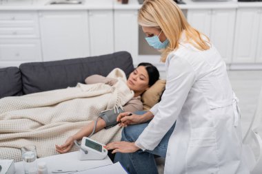 Tıp maskeli doktor evde bulanık Asyalı bir hastanın yanında tonometreye bakıyor. 