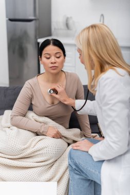 Middle aged doctor with stethoscope checking lungs of asian patient on couch at home  clipart