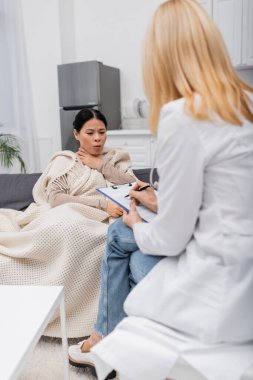 Sick asian woman coughing near blurred doctor writing on clipboard  clipart